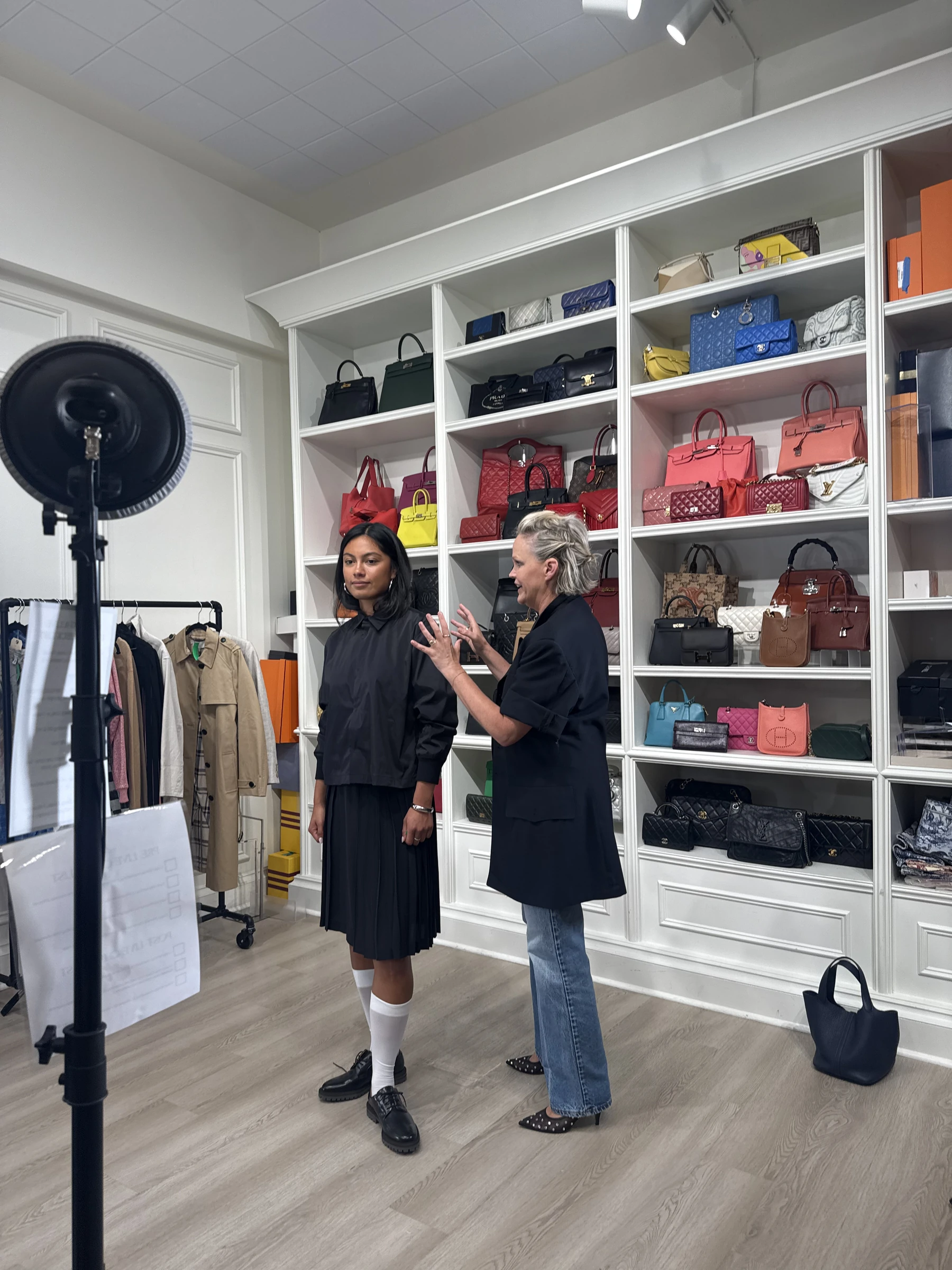A stylist adjusts a model's outfit beside a livestream ring light and styling racks.